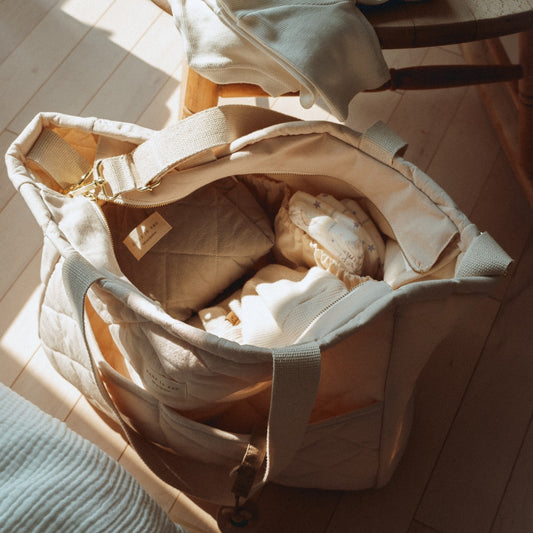 An open beige cotton quilted diaper bag with beige straps placed on a floor, made in Quebec, Canada.
