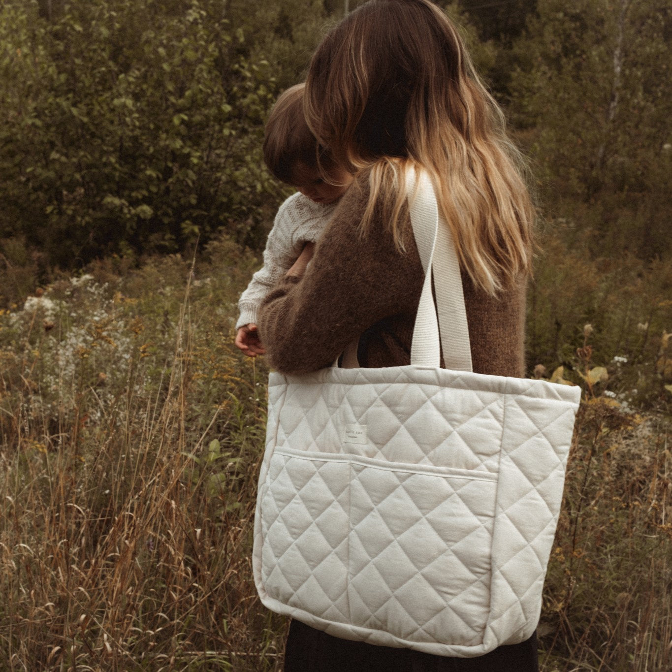 A mother carrying her baby and the beige quilted cotton diaper bag, made in Quebec, Canada.