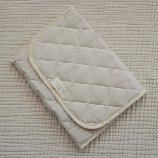 A folded beige cotton quilted baby changing mat with a visible brand label, 100% cotton, made in Quebec, Canada.