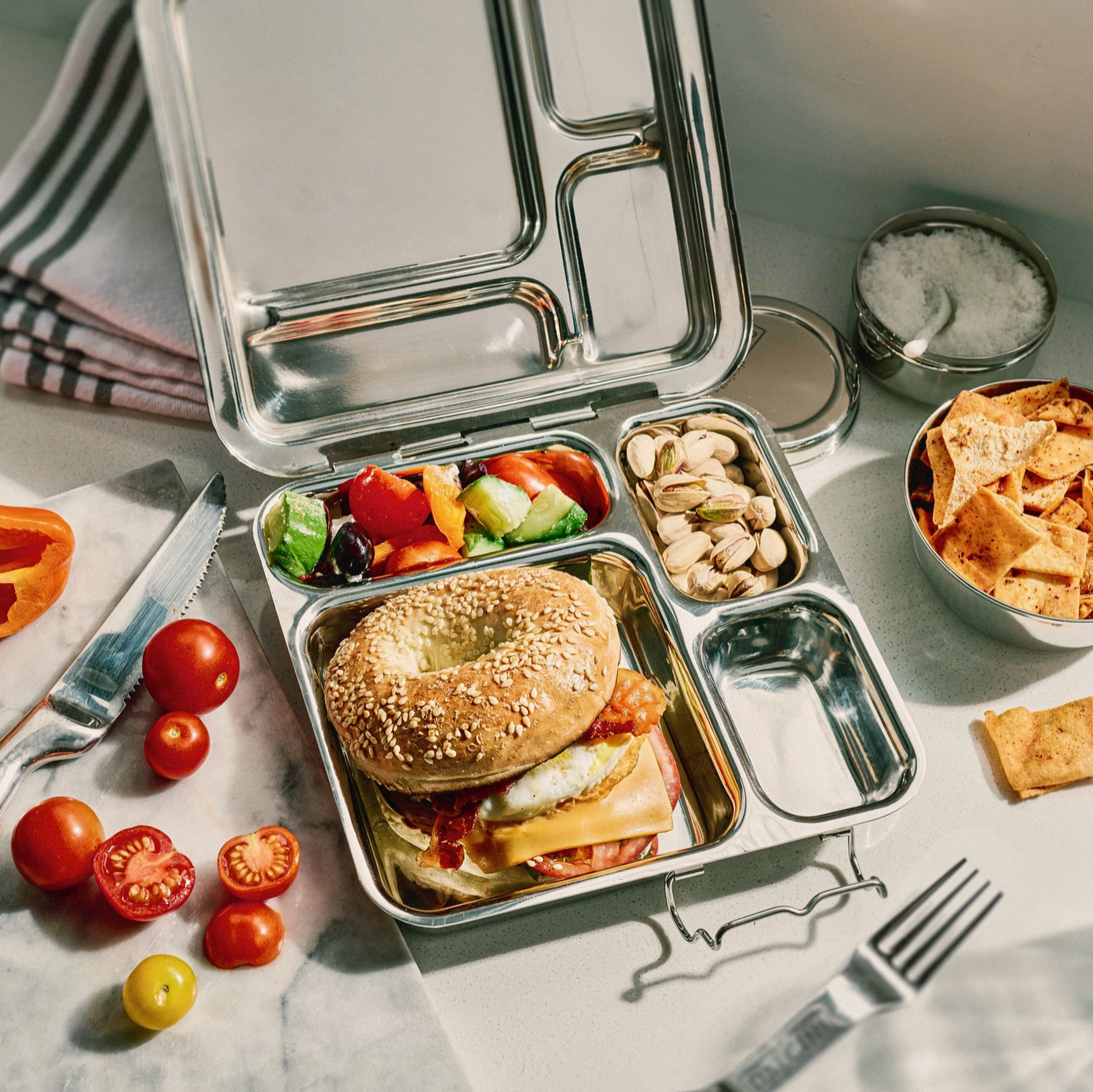 A stainless steel bento box lunch with various compartments containing food items like a bagel, fruits, nuts, and some chips, placed on a table with a few cherry tomatoes and a napkin visible.