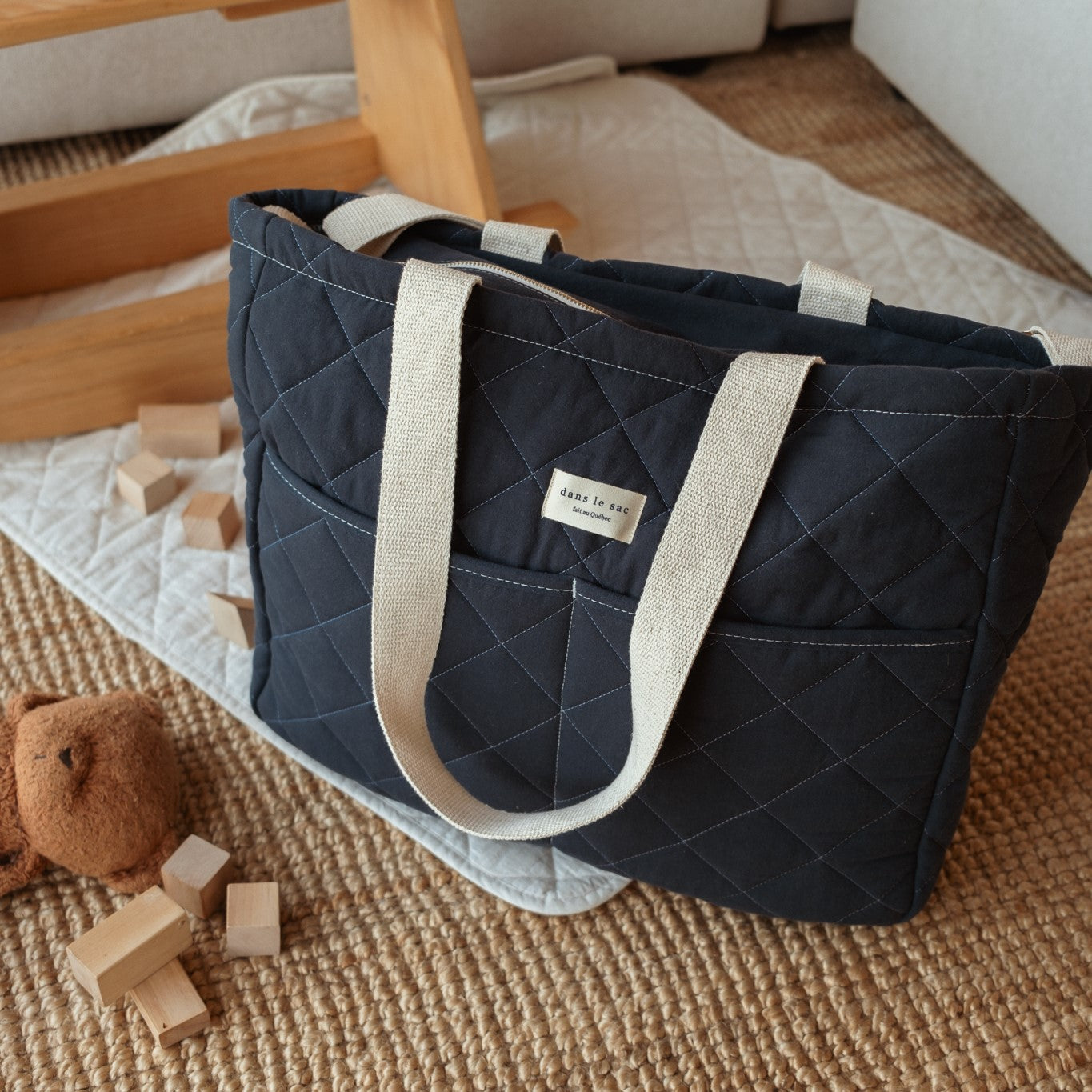 A navy blue cotton quilted diaper bag with beige straps placed on a floor next to a wooden toy and some blocks. Made in Quebec, Canada.